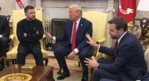 Wolodymyr Selenskyj, Donald Trump und JD Vance im Oval Office. Selenskyj hat die Arme verschränkt, Trump und Vance strecken ihm abwehrende Handflächen entgegen, Vance hat das Gesicht leicht abgeneigt.