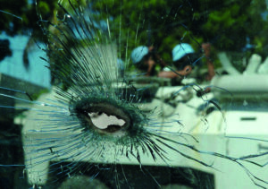 Im Vordergrund Loch und Splitter in einer Glasscheibe, im Hintergrund ein Tankwagen der UN mit UN-Soldaten, der im Viertel Bel-Air von Anhängern Aristides angegriffen wird.