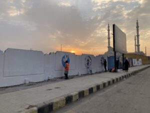 In the foreground, a woman wearing an orange vest is painting a dove of peace on a white wall. Other people nearby are creating murals, including another dove and abstract designs. The scene takes place on a wide street in Aleppo, with two minarets and a sunset visible in the background. The sky is cloudy and the lighting is subdued. A large dark sign stands to the right of the murals.