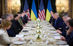 Several people are seated at a set table. Among them are Zelenskyy and Trump. In the background, two U.S. flags and two Ukrainian flags can be seen.