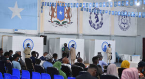 Room filled with people sitting on plastic chairs, with blue flags hanging from the ceiling. On the wall there are symbols of the Somali Police Force, and the Somali flag. Security guards stand in front of booths.