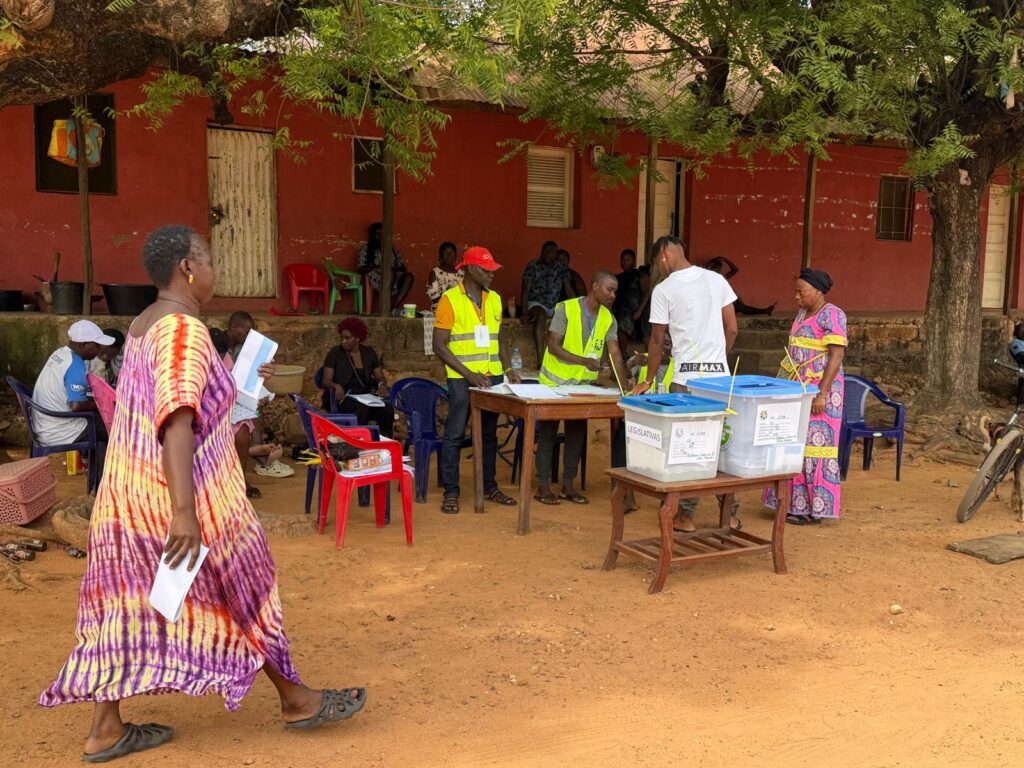 Voting scene: A table with election officers, ballots. Two women and one man are submitting thir vote to the ballots. 